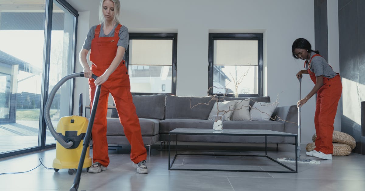 Two women in red overalls vacuuming and sweeping a modern living room.