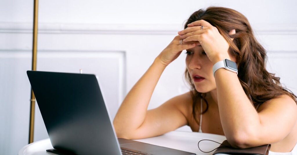 Woman feeling stressed while working on a laptop, using smartphone and smartwatch at a desk.
